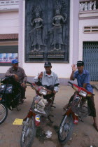 Three motorbike taxi drivers on their mopeds beneath a large wall plaque depicting AsparasAsian Cambodian Kampuchea Southeast Asia 3 Kamphuchea