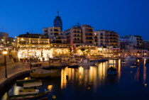 Saint Julians Bay waterfront at dusk with illuminated restaurants in front of apartments and the Hilton Tower office complex with fishing boats in the harbour in the foreground