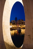 Saint Julians Bay waterfront at dusk with illuminated restaurants in front of apartments and the Hilton Tower office complex with fishing boats in the harbour in the foreground seen through a spherica...