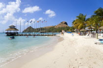 Tourists on the beach at Sandals Grande St Lucian Spa and Beach Resort hotel beside a wooden jetty with Pigeon Island National Historic Park beyond