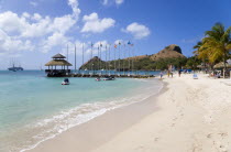 Tourists on the beach at Sandals Grande St Lucian Spa and Beach Resort hotel beside a wooden jetty with Pigeon Island National Historic Park beyond