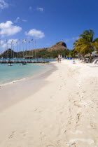 Tourists on the beach at Sandals Grande St Lucian Spa and Beach Resort hotel beside a wooden jetty with Pigeon Island National Historic Park beyond