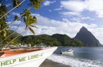 Fishing boats on the beach lined with coconut palm trees with the town and the volcanic plug mountain of Petit Piton beyond. A fishing boat in the foreground with the words Help Me Lord written on the...