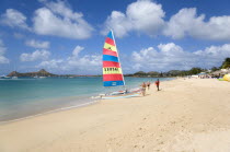 Reduit Beach in Rodney Bay with tourists on the beach beside a hobbycat catamarran with yachts at anchor in the bay and Pigeon Island in the distance