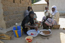 Man sitting in shade in yard outside his house in the company of two of his wives who are taking care of a child while washing and smashing tomatoes to be used in the familys meal TanjehTanjihhouse...