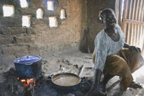 Young woman cooking a traditional Gambian dish of groundnut sauce called mafay over open fire in kitchen situated outside  house with sunlight filtering through small  square openings in the wall behi...
