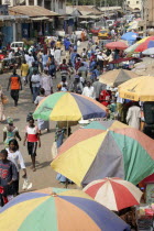 Bakau Market  Atlantic Road.  Busy city street lined with stalls beneath colourful striped sun umbrellas.  Crowds of people  traffic and shop fronts open to the street.                   SerecundaSe...