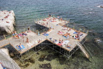 Looking down on platform structure raised above rocky sea with steps to water  people sunbathing  playing and swimming.Siracusadiving-boardjettypierSummerholidayvacationEuropean Holidaymakers...