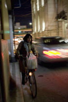 Gion District.  Japanese woman riding her bicycle at night along a side road in Kyoto carrying bag of shopping in basket on the handlebars with car passing in the opposite direction.vehicletransport...