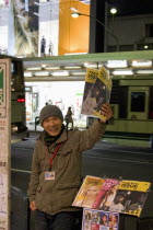 Japanese man selling the  Big Issue  magazine  arm raised  holding copy with passing traffic on street behind and lighted shop windows in evening.modern lifehomelessunemployedunemploymentjob seek...