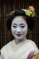 Gion District.  Head and shoulders portrait of smiling apprentice Geisha or Maiko with hair pinned up with flowers and decorative ornaments  white facial  make up  red painted lips  and wearing tradit...
