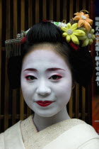 Gion District.  Head and shoulders portrait of smiling Maiko or apprentice Geisha  with hair worn up and fixed with decorative pins and flower ornaments  white facial make up and red painted lips wear...