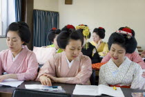 Gion District.  Geisha and Maiko apprentice Geisha attending a class at Mia Garatso school of Geisha.  Three young women wearing plain kimono seated at desk in foreground with open books.Geikotradit...
