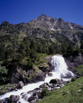 Vallee du Lutour.  Jagged peaks of Soum det Guingays 2451 m / 8027 ft above white waterfall.  Outcrops of rock scattered over hillside amongst trees.