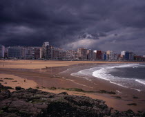 High rise city buildings overlooking beach with people in water  on beach and surfing.  Grey  cloudy sky with approaching storm.  Rocks and seaweed in foreground.