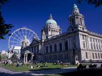 People sitting or lying in the sun in the grounds of City Hall with The Wheel of Belfast part seen behind.ferris wheel Bal Feirste Eire European Irish Northern Europe Republic Ireland Poblacht na h...