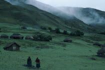 Village thatched homes on green hillside with mist seen high above. Two women standing together at the bottom of the slope 2 American Female Woman Girl Lady Hispanic Latin America Latino Peruvian Sce...