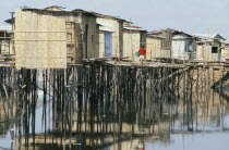 Slum housing with stilt buildings built over untreated sewage. A woman wearing a red cardigan carrying a bucket along walkway with reflections on the water belowAmerican Equador Female Women Girl Lad...