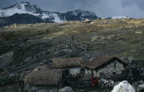 Collpa Huata Llama herders settlement of stone houses with thatched roofs. A woman wearing traditional red textiles standing next to stone wall. Snow covered mountains behind. Near PeruAmerican Boliv...