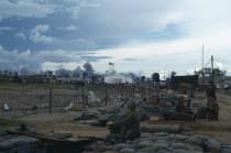 Vietnam War. Siege of Kontum. American B-52 bombing behind Kontum. Soldiers sitting in camp within a barbed wire perimeter watching plumes of grey smoke rising into the sky Kon TumAsian Southeast As...