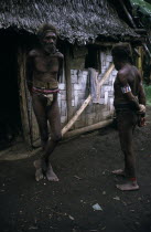 Two men wearing penis sheaths standing outside thatched hut.Pacific islands 2 Indegent Male Man Guy Old Senior Aged