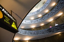 Vatican City Museums Tourists descending the Spiral Ramp designed by Giuseppe Momo in 1932 leading from the museums to the street level below seen from below with an illuminated sign warning to watch...