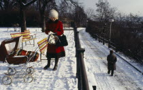 Woman stopped to read newspaper in park in the snow.  She wears winter coat  hat and boats with baby in push chair beside her covered with thick blanket.Babies Ceska Eastern Europe European Female Wo...