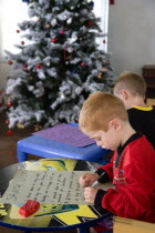 Two young left handed boys on Christmas Eve sitting down on a sofa and writing their Santa Lists at tables in front of the Christmas TreeCultural Cultures Immature Kids Order Fellowship Guild Club Re...
