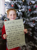 Young smiling red headed boy standing in front of a decorated Christmas Tree holding up the Santa List he has written on Christmas EveCultural Cultures Immature Kids Order Fellowship Guild Club Relig...