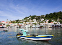 Fishing boat moored in the Carenage harbour of the capital city of St Georges with houses and the roofless cathedral damaged in Hurricane Ivan on the nearby hill. A boy in a speedboat heads inshore pa...