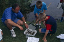 Pond Dipping activity. Children with adult inspecting a fishing net to identify species of aquatic life from worksheets in ecological study . Norfolk  England.Outing Teacher European Great Britain Im...