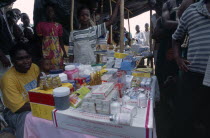 Woman behind stall with display of medical supplies surrounded by crowd of adults and children.    African Female Women Girl Lady Kids Liberian Western Africa Female Woman Girl Lady Libirian