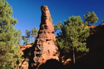 A tall thin  eroded rock pinnacle in the area known as the Needle Cirque part circled by trees against cloudless blue sky.Ochre Trail European French Western Europe