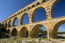 Pont du Gard.  Angled view of Roman aqueduct from the west side in glowing evening light showing three tiers of continuous arches against cloudless blue sky.Bridge arch stone European French Western...