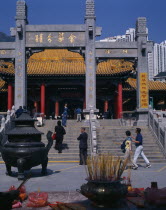 Wong Tai Sin taoist temple established in 1921.  Visitors on flight of steps to entrance built in traditional Chinese style with red pillars and pagoda style roof.  Incense  offerings and burner in f...