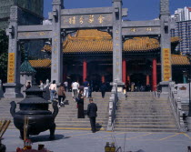 Wong Tai Sin taoist temple established in 1921.  Visitors on flight of steps to entrance built in traditional Chinese style with red interior pillars and pagoda style roof.  Incense  offerings and bu...