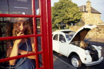 Girl making a call in a traditional red phone box with broken down Citroen 2CV car outside Automobile Automotive Cars Classic Classical European Historical Motorcar Older One individual Solo Lone Sol...