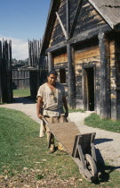 Sainte Marie among the Hurons near Midland. Indian man pushing a wheel barrel near wooden building.American Canadian Indegent Male Men Guy North America Northern One individual Solo Lone Solitary 1 M...