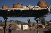 Family enclosure with circular hut beside frame of roof  passing woman and raised platform in foreground with sacks and baskets on top.African American Female Women Girl Lady Senegalese Southern Fema...