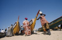 Women wearing brightly coloured traditionally patterned clothing on beach with men and children standing besidepainted fishing boats.Beaches Colored Female Woman Girl Lady Kids Male Man Guy Resort Sa...