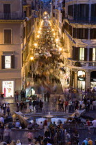 The Via dei Condotti the main shopping street busy with people illuminated at night seen from the Spanish Steps with seated tourists and the Fontana della Barcaccia in the foregroundEuropean Italia I...