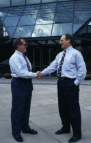 Two businessmen in conversation shaking hands outside modern glass fronted building in downtown business area.talking agreement gesture 2 American North America United States of America Northern