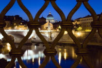 The Dome of the Basilica of St Peter in the Vatican City illuminated at night with the Ponte Vittorio Emmanuel II bridge across the River Tiber in the foreground all seen through iron railings on the...