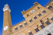 The Torre del Mangia campanile belltower and facade of the Palazzo Publico town hall in the Piazza del Campo under a blue sky. The black and white crest or symbol of Siena decorates the facade of the...