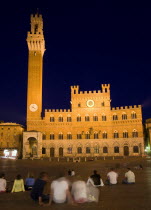 The Torre del Mangia campanile belltower and facade of the Palazzo Publico town hall in the Piazza del Campo and surrounding buildings illuminated at night with tourists sitting on the ground in the s...