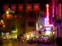 Via Santa Maria leading off the south side of the Campo dei Miracoli or Field of Miracles. People sitting at outdoor restaurant tables at night lining the pavement. Brightly coloured neon sighs advert...