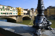 The 14th Century Ponte Vecchio bridge across the River Arno showing the backs of the goldsmiths workshops that hang over the water with an ornate wrought iron lamp post with feet in the foregroundEur...