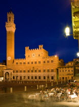 The Torre del Mangia campanile belltower and facade of the Palazzo Publico town hall in the Piazza del Campo illuminated at niight with people seated at tables in the square outside a restaurant Euro...