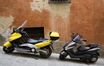 Two moped scooters one yellow one grey parked on a sloping sidestreet beside a red ochre wallEuropean Italia Italian Southern Europe Toscana Tuscan 1 2 Gray Single unitary