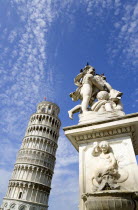 The Campo dei Miracoli or Field of Miracles.The Leaning Tower or Torre Pendente belltower with tourists on the top level under a blue sky with a statue and water fountain with cherubs in the foregroun...